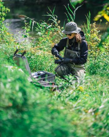 A person kneeling in a grass near a river with measuring tools